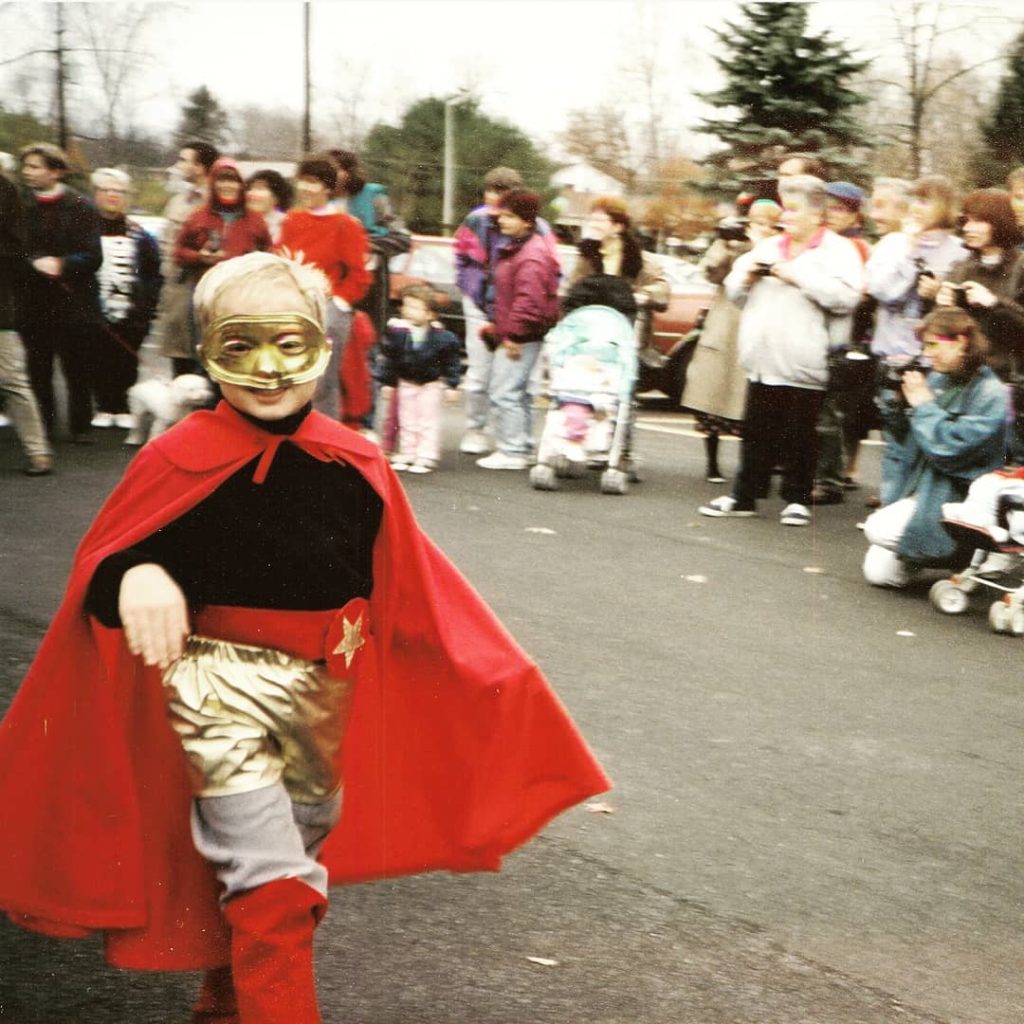 Photo of the artist from childhood wearing a homemade super hero costume including gold lamé shorts and a red cape, swishing a limp wrist.